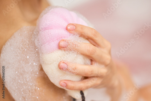 Woman's hand holding a pink bath sponge, scrubbing her skin and creating abundant white soap bubbles during a relaxing bath for hygiene