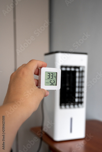 Hand holding a digital room thermometer showing high temperature of 33°C against a blurry portable air cooler background. Summer heat and climate control concept