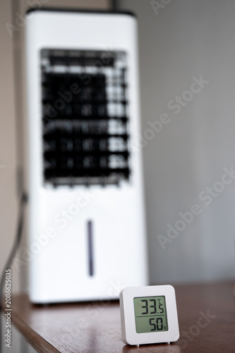 A digital room thermometer displaying a high temperature of 33°C, set against the blurred background of a portable air cooler. Summer heat and the concept of climate control