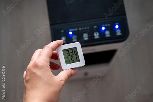 Hand holding a digital room thermometer showing high temperature of 33°C against a blurry portable air cooler background. Summer heat and climate control concept