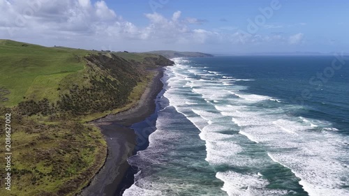 Wide aerial view following the dramatic black sand beach of New Zealand's North Island, where rolling ocean waves meet steep coastal hills in a spectacular natural seascape.