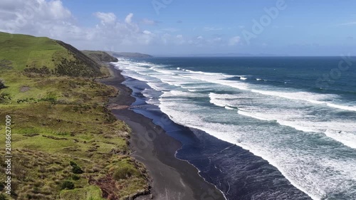 Sweeping aerial panorama of North Island, New Zealand, featuring a rugged black sand shoreline, rolling surf, and pristine Pacific coastline under clear blue skies.