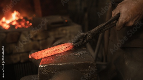 Blacksmith holding a glowing hot metal bar with tongs on an anvil in a workshop. Traditional medieval craftsman forging iron. Industrial manufacturing of metal tools and steel items.