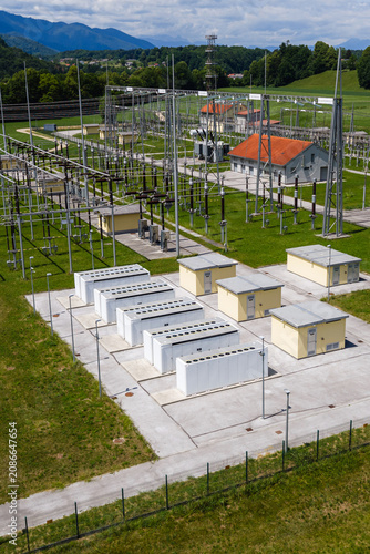 Aerial view shows battery energy storage system near power lines and buildings with green fields and mountains in the background.