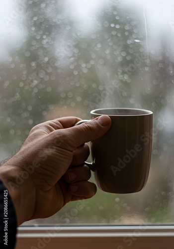 Hand Holding Hot Coffee Mug Next to a Rainy Window Creating a Cozy Atmosphere of Relaxation and Contemplation Indoors on a Cloudy Day