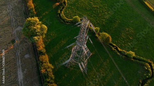 High voltage transmission tower above agricultural landscape at golden hour. Aerial view with power lines crop fields green meadows and rural infrastructure. Electricity distribution network energy