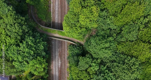 Aerial drone footage looking directly down over a pedestrian footbridge crossing railway tracks surrounded by dense woodland in Leeds West Yorkshire England showing transport infrastructure and trees