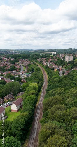 Portrait vertical aerial drone footage looking along a railway line through suburban Leeds West Yorkshire England with residential housing mature woodland and transport infrastructure