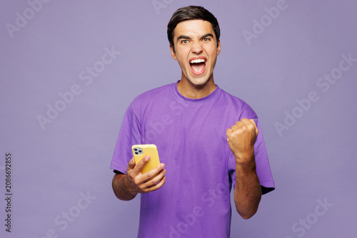 Young happy Caucasian man he wear violet t-shirt casual clothes hold in hand use mobile cell phone do winner gesture isolated on plain pastel light purple background studio portrait Lifestyle concept