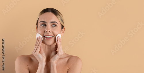 Cuadro en lienzo Young beautiful woman smiling while using cotton pads on her cheeks in a beige s