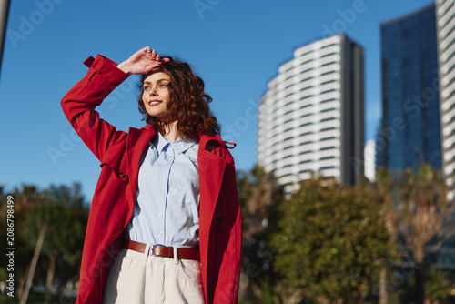 Fotografía Young woman with curly hair wearing red coat and white shirt standing outdoors in city park with modern buildings in background on sunny day