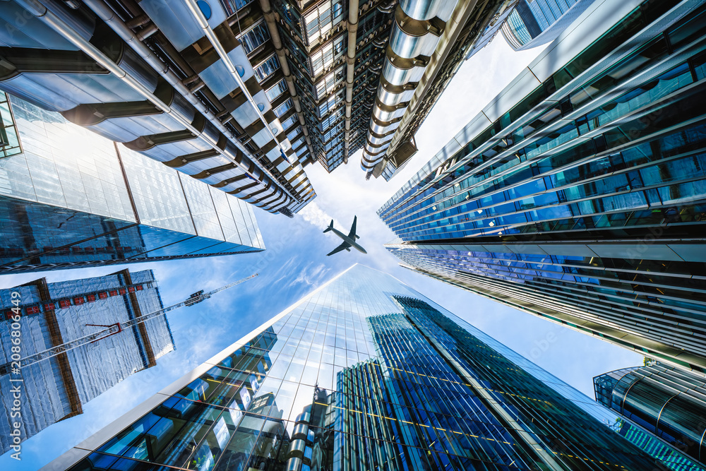 Naklejka premium Low angle view of looking up directly at the skyline of the financial district in central London with an airplane in the sky passing by