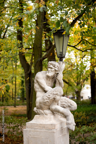 Bild auf Leinwand Ancient mythological faun sculpture built as a street light post amidst colorful autumn trees with yellow and green leaves