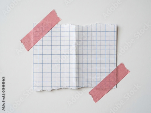 Torn Piece Of White Grid Paper With Blue Lines Secured By Two Pink Decorative Tape Strips On White Wall