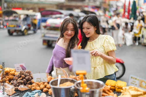Two asian women buying deep fried street food at local Thai market  Authentic travel experience  culinary tourism  summer vacation destination  outdoor night market and joyful food tasting lifestyle