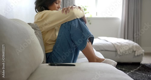 African American woman sitting on light sofa at home, phone causing curling and lowering head