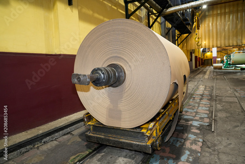Massive industrial paper roll on a yellow wheeled trolley sits on a concrete floor, ready for transport within a paper mill production area filled with machinery