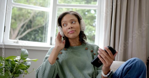 African American adult woman with headphones tapping smartphone checking song and singing by window