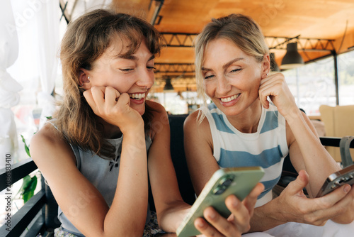 Two women of different ages are sitting in a café, scrolling through a social media feed on a phone.