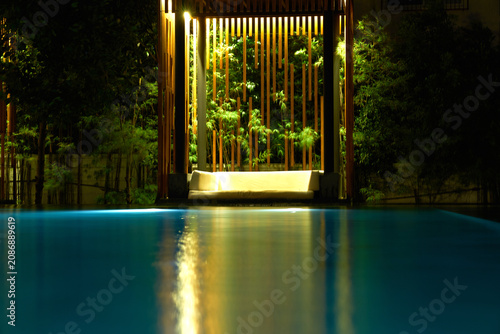 A luxury swimming pool at night reflecting a warm golden lit wooden pavilion structure with vertical bamboo or timber slats.
