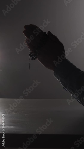 Vertical video person silently praying with folded hands and a rosary, symbolizing faith, devotion, and spiritual reflection during a moment of solemn contemplation in a dimly lit setting