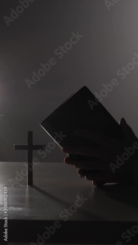 Vertical video person holding a bible, forming a silhouette against a dark background, symbolizing faith, devotion, prayer, and spirituality, with a wooden cross reflecting religious belief