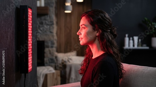 Woman sitting in front of red light therapy device. Wellness treatment for skin rejuvenation and recovery. Modern home healthcare technology for healing and beauty skin care routine.