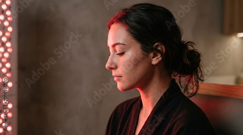 Woman sitting in front of red light therapy panel for skin rejuvenation and wellness. Photobiomodulation treatment for anti aging, recovery and relaxation in meditative home spa setting.