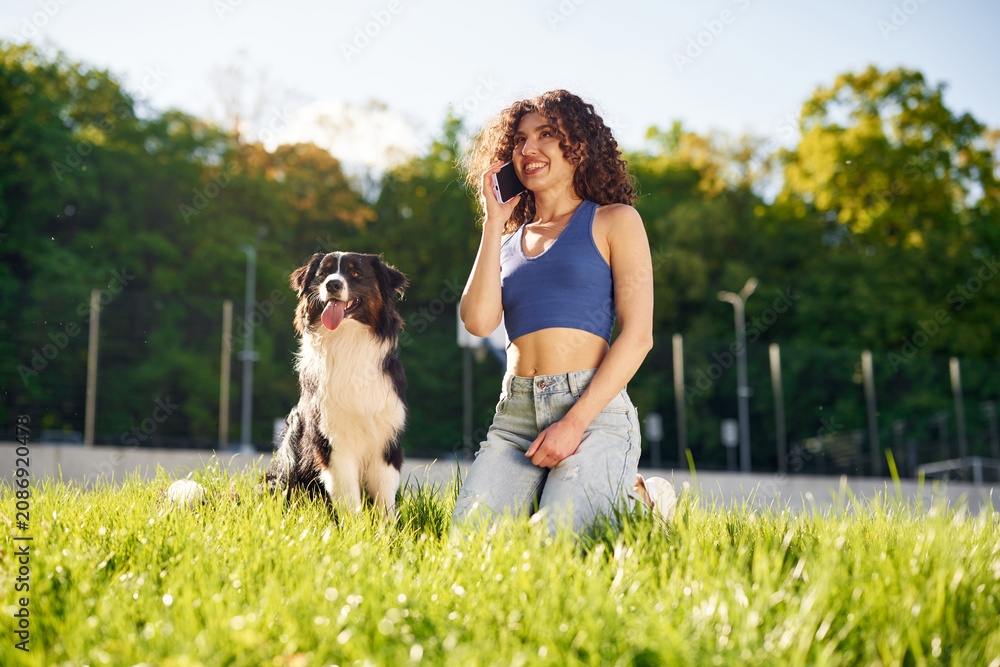 Fototapeta premium Young woman with curly hair kneeling on grass while talking on smartphone beside a black and white dog in a sunny park setting with trees in the background