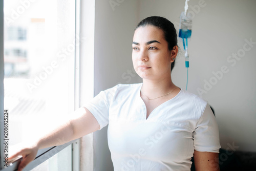 Female nurse standing beside an IV drip while looking through a hospital window in a bright healthcare environment.