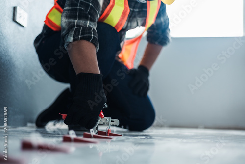 Obraz na płótnie A worker in a safety vest and black gloves uses a tile leveling plier tool to adjust red wedges on a floor, installation and professional interior renovation