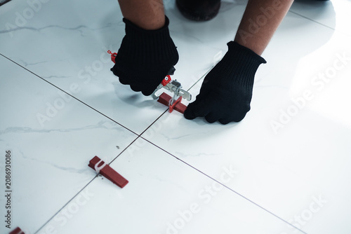 Fotografie A worker in a safety vest and black gloves uses a tile leveling plier tool to adjust red wedges on a floor, installation and professional interior renovation