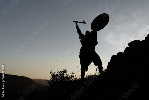 Foto Silhouette of a warrior stands triumphantly against a dusky sky, holding aloft a shield and sword