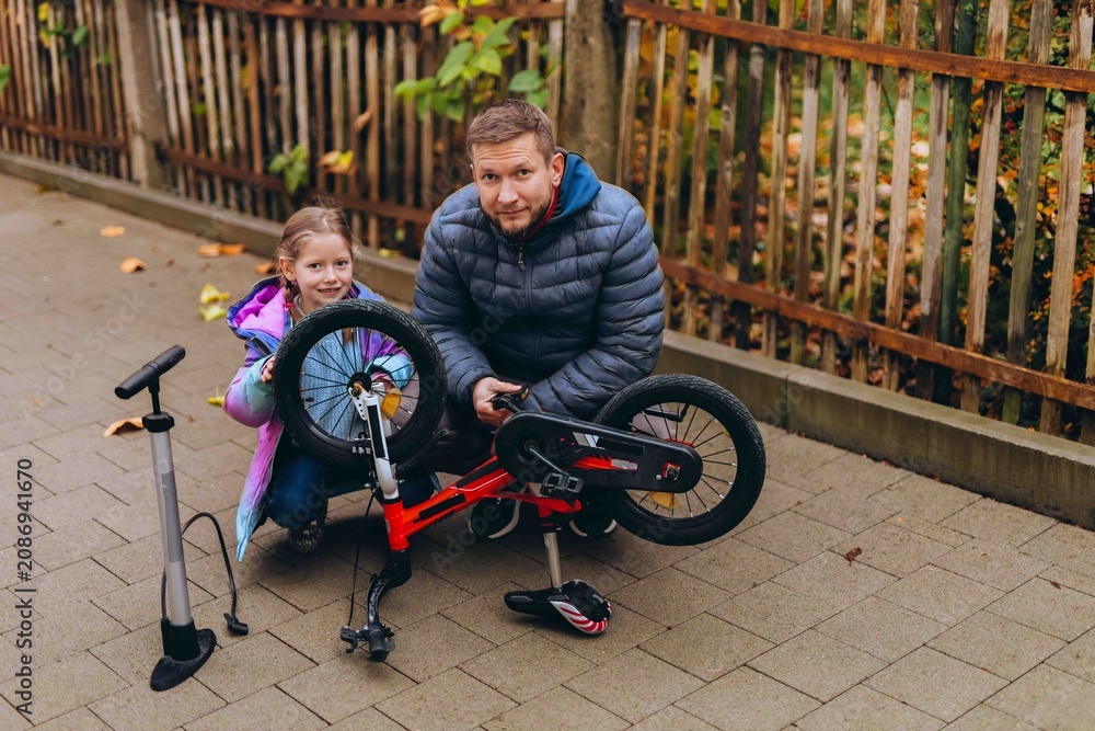 Obraz premium Father and daughter examine an upside down balance bike together before a ride. Shared bicycle maintenance turns everyday repairs into practical family learning.