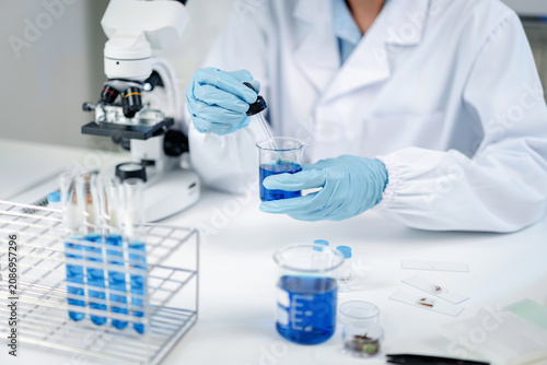 Scientist in white lab coat and blue gloves using dropper pipette to add solution into beaker of blue liquid, with microscope, test tubes and specimen slides on laboratory table