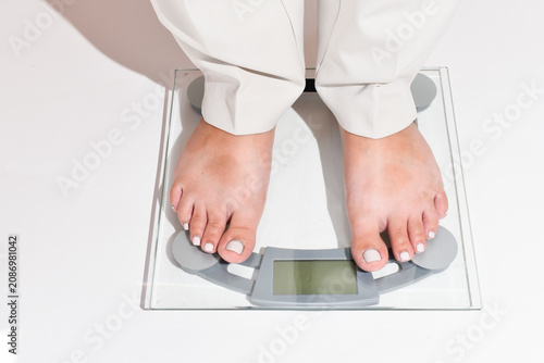 Woman’s legs in white pants step onto a bathroom scale on a clean white background. Close-up of feet with a light gel pedicure standing on digital scales. Weight control, weight loss journey