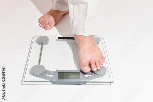 Woman’s legs in white pants step onto a bathroom scale on a clean white background. Close-up of feet with a light gel pedicure standing on digital scales. Weight control, weight loss journey