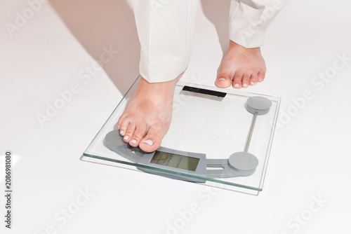 Woman’s legs in white pants step onto a bathroom scale on a clean white background. Close-up of feet with a light gel pedicure standing on digital scales. Weight control, weight loss journey