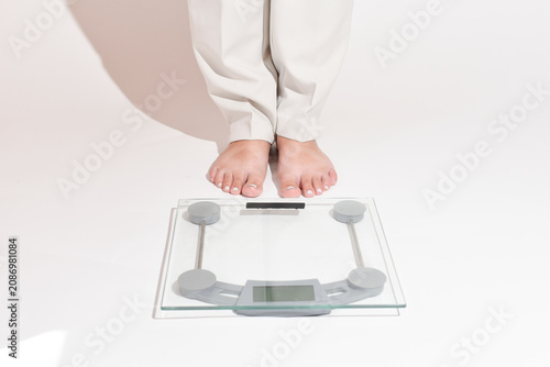 Woman’s legs in white pants step onto a bathroom scale on a clean white background. Close-up of feet with a light gel pedicure standing on digital scales. Weight control, weight loss journey