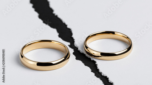 Two gold wedding rings separated by a torn sheet of white paper over a black background, symbolizing divorce, marital separation, and the end of a relationship.