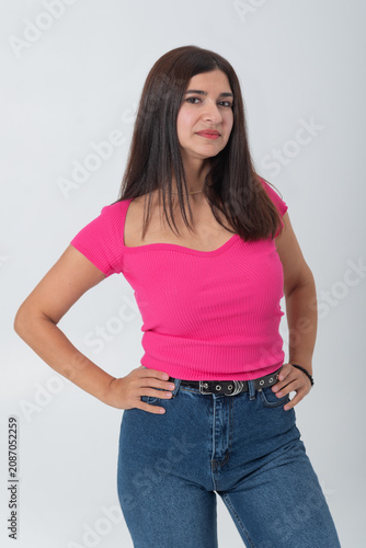 Confident young woman standing in a studio against a white background, hands on hips, wearing a vibrant pink top and blue denim jeans, looking directly at camera with serious poise