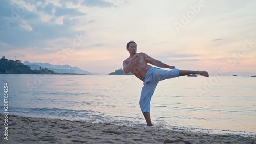 Shirtless man practicing martial arts on sandy shoreline at dusk with raised fists, high side kick, balanced stances, gentle waves, pastel sky, and distant mountains