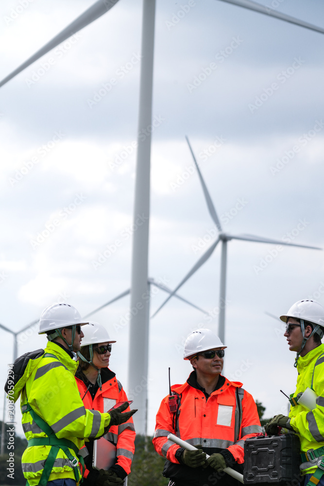 Fototapeta premium Wind project engineers with laptop, radio and drawings swap the laptop between them to verify a figure gathered on open ground beneath the turbines in silver overcast light on a working.