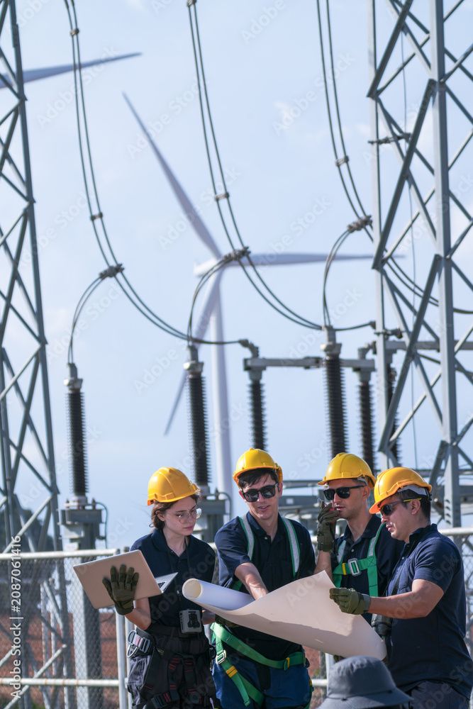 Fototapeta premium A commissioning team in navy suits and yellow hard hats check disconnector positions against the diagram inside the wind farm grid substation in crisp blue sky daylight at an operating.