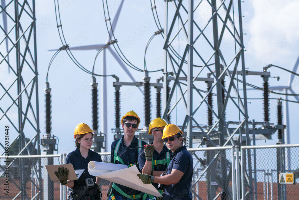 Fototapeta premium A commissioning team in navy suits and yellow hard hats check disconnector positions against the diagram inside the wind farm grid substation in crisp blue sky daylight deep inside a.