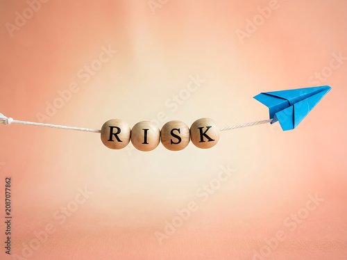 Blue paper airplane flies past wooden beads spelling RISK on a tightrope during soft studio lighting