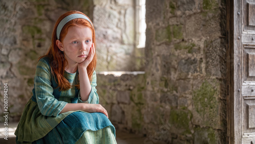 A pensive young girl with red hair and a wistful expression in a medieval style dress sits and poses within a mossy stone wall.
