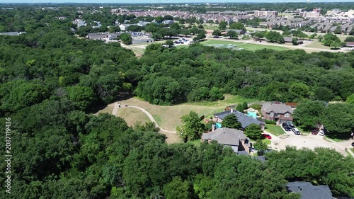 Concrete recreation paths weave through grassy clearings adjacent to municipal forest land. Mature trees stand against blue sky, marking natural community boundaries in Flower Mound, Texas