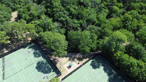 Tennis courts border dense woodland near Leonard and Helen Johns Community Park. Green hard courts contrast with vibrant summer foliage bright sun, suburban recreation in Flower Mound, Texas