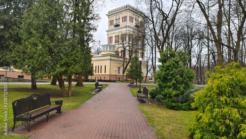 The palace with arched windows has a clock tower. In front of him, the park's trees grow on grassy lawns, and benches and lampposts line the tiled paths. Cloudy spring weather and grey sky